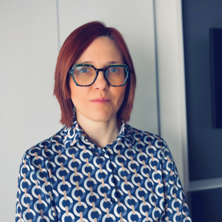 Portrait of a woman with short red hair and black framed glasses wearing a blue patterned blouse standing indoors